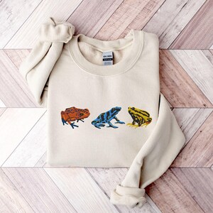 May include: A cream-colored sweatshirt with a graphic of three frogs in different colors: orange, blue, and yellow. The frogs are facing forward and have black eyes.