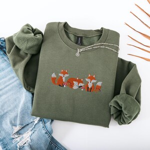 May include: A green sweatshirt with an embroidered design of three foxes in a forest setting. The foxes are orange and white with black eyes and noses. The design includes small brown mushrooms and green leaves.