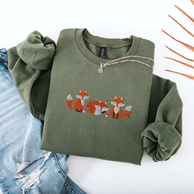 May include: A green sweatshirt with an embroidered design of three foxes in a forest setting. The foxes are orange and white with black eyes and noses. The design includes small brown mushrooms and green leaves.