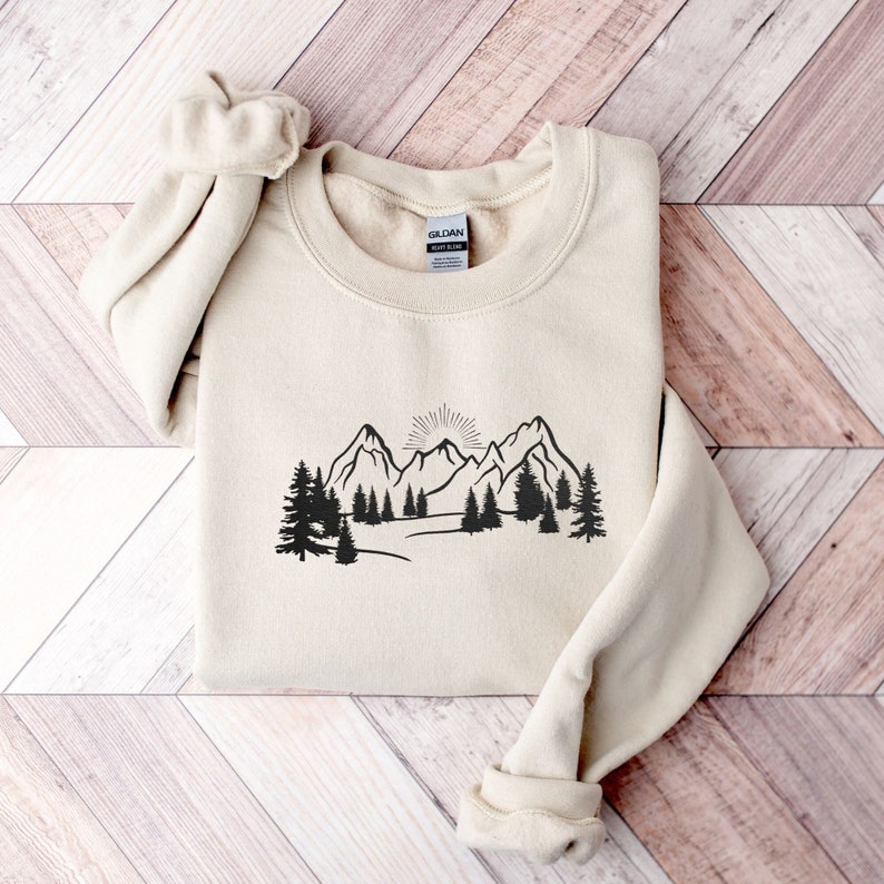 Op de afbeelding: Een beige sweatshirt met een zwarte afbeelding van een bergketen met een opkomende zon erachter. De bergen zijn omringd door bomen.