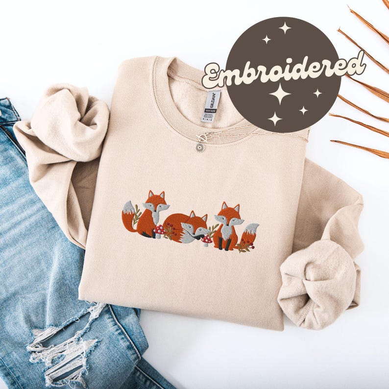 May include: A beige sweatshirt with an embroidered design of three foxes. The foxes are orange and have black eyes and noses. The design includes red mushrooms and green leaves.