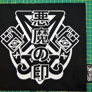 May include: Black and white patch with a graphic design featuring a stylized eye, a triangle, and Japanese text that reads "The Demon's Eye". The patch is sewn onto a black fabric background.