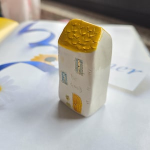 Könnte beinhalten: Kleine, weiße Keramikhausfigur mit gelbem Dach, blauen Fensterrahmen und einer gelben Tür. Auf der Vorderseite steht der Schriftzug "be good". Das Haus steht auf einer weißen Oberfläche.