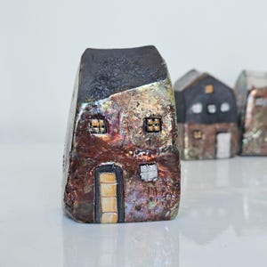 May include: A ceramic house sculpture with a textured, metallic finish in shades of brown, copper, and black. The house features two small windows and a door with a yellow glow. Other houses are slightly out of focus in the background.
