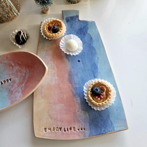 Könnte beinhalten: Eine Keramik-Servierplatte mit einem Aquarell-Design in Blau, Rosa und Gelb. Die Platte hat eine rechteckige Form mit abgerundeten Kanten und einen kleinen Griff oben. Die Platte trägt die Aufschrift "Enjoy Life..." auf der Unterseite.