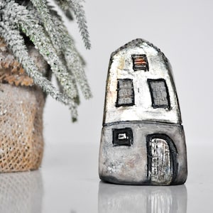 May include: A small, gray and silver house figurine with a black door and windows. The house is made of clay and has a rustic, handmade look.