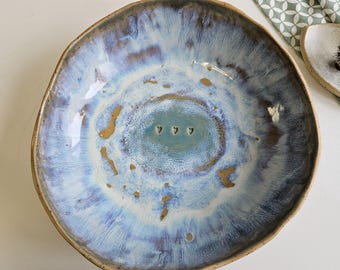May include: A handmade ceramic bowl with an irregular shape. It has a glossy glaze in blue, white, and brown hues. The centre has three small heart-shaped cutouts. The rim is a darker brown. A decorative piece for the home.