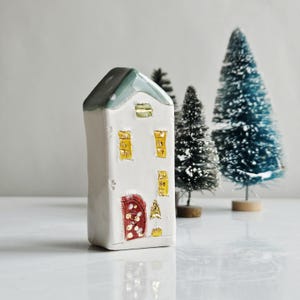 Può includere: Una casa in ceramica bianca con tetto verde, finestre gialle e porta rossa. La casa è decorata con piccoli dettagli e si trova di fronte a diversi alberi di Natale in miniatura. Un'affascinante decorazione natalizia.