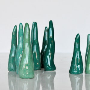 Puede incluir: Un grupo de esculturas de cerámica de color verde azulado con forma de árboles altos y delgados. Las esculturas están dispuestas en fila, con algunas que se superponen ligeramente. Las esculturas son brillantes y tienen una superficie lisa y redondeada.