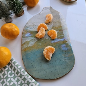 Puede incluir: Una fuente de cerámica hecha a mano con una forma orgánica única. La fuente presenta una mezcla de esmalte azul, verde y marrón, salpicado de puntos oscuros. Varios segmentos de mandarina pelados están dispuestos en la fuente. La escena se desarrolla sobre una superficie blanca.