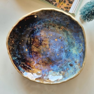 Könnte beinhalten: Eine blaue und braune Keramikschale mit einem gesprenkelten Überzug und goldenen Sternen. Die Schale hat eine leicht unregelmäßige Form und eine glänzende Oberfläche.