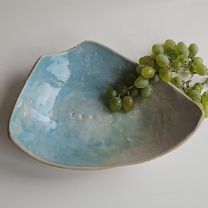 May include: A handmade ceramic serving bowl with a light blue and gray glaze. The bowl has a unique, asymmetrical shape and three heart-shaped cutouts. A bunch of green grapes rests in the bowl.