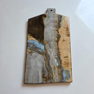 Könnte beinhalten: Ein rechteckiges Servierbrett aus Keramik mit spitz zulaufendem Ende und einem kleinen Aufhängeloch. Die Oberfläche zeigt ein abstraktes Glasurmuster in Dunkelbraun, Creme, Beige und Hellblau, was für eine strukturierte Optik sorgt.