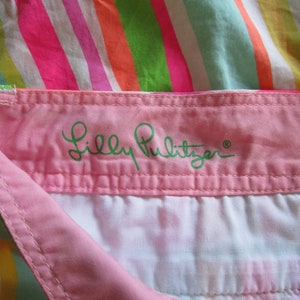 May include: Close-up of a colorful striped fabric with a pink waistband. The waistband features the embroidered brand name "Lilly Pulitzer" in green script. The stripes include pink, orange, green, and white.