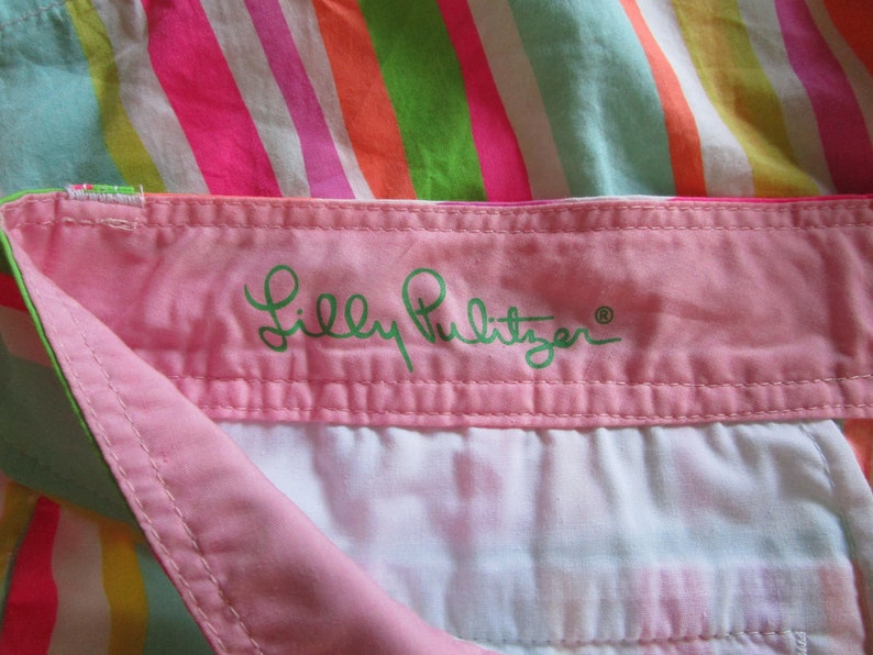 May include: Close-up of a colorful striped fabric with a pink waistband. The waistband features the embroidered brand name "Lilly Pulitzer" in green script. The stripes include pink, orange, green, and white.