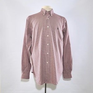 May include: A long-sleeve button-down shirt featuring a small-scale red and white gingham check pattern. The shirt has a button-down collar, a front button placket, and a small dark embroidered logo on the left chest area. The cuffs are buttoned.