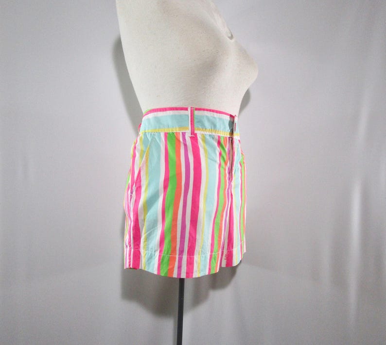 May include: A pair of colorful striped shorts in shades of pink, green, orange, and blue. The shorts have a waistband with a button closure and belt loops. The garment is displayed on a mannequin.