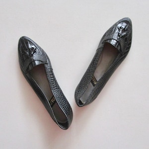 May include: A pair of black leather loafers with a textured, reptile-like pattern. Each shoe features a decorative tassel and a low heel. The shoes are displayed on a light-colored surface.