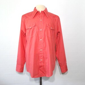May include: A coral-colored button-down shirt with long sleeves, two chest pockets, and a pointed collar. The shirt features white stitching and white buttons. The shirt is displayed on a mannequin.