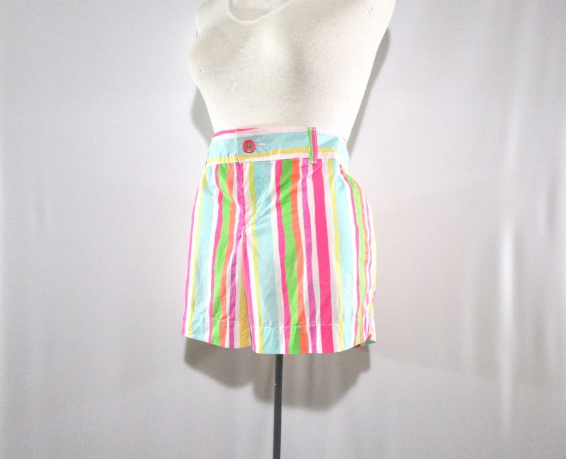 May include: A pair of colorful striped shorts in a variety of bright colors, including pink, green, blue, and yellow. The shorts have a button and a belt loop. The garment is displayed on a mannequin.