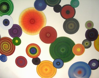 May include: A white wall with a colorful display of round, painted discs. The discs are various sizes and feature different patterns, including concentric circles, radial lines, and abstract designs. The colors include green, red, orange, blue, purple, yellow, and black.