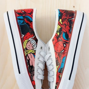 May include: A pair of white canvas shoes with a comic book print featuring Spiderman, Thor, and other superheroes. The shoes have black laces and white soles.