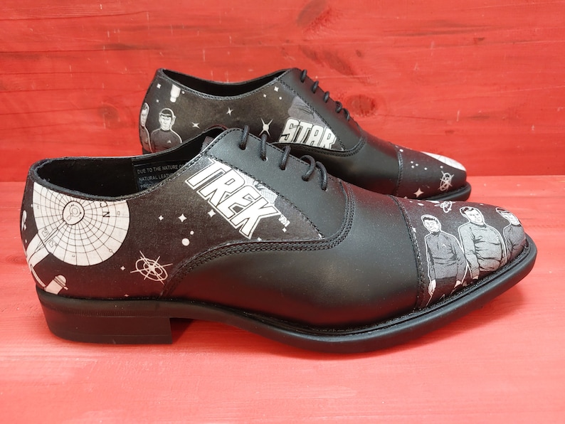 May include: Black leather lace-up shoes with a white and black space-themed print. The shoes feature the words "Star Trek" and a depiction of the Starship Enterprise.