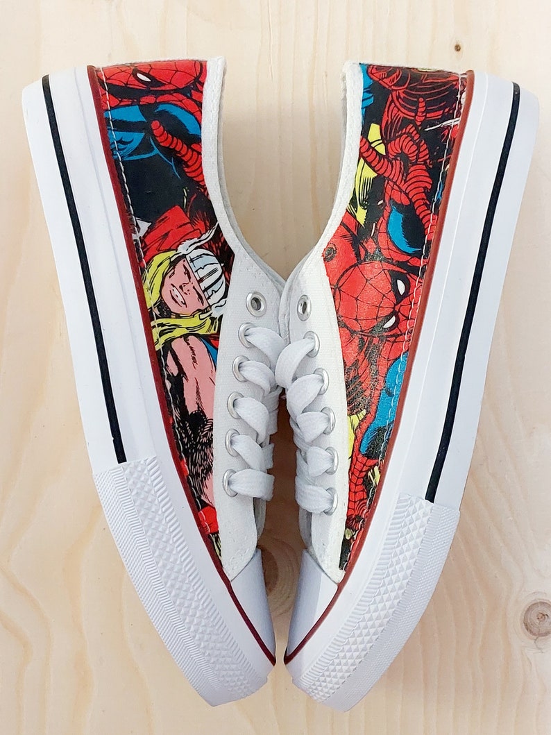May include: A pair of white canvas shoes with a comic book print featuring Spiderman, Thor, and other superheroes. The shoes have black laces and white soles.