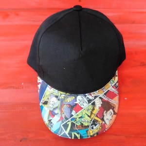 May include: Black baseball cap with a comic book print on the brim. The print features a variety of characters and scenes.