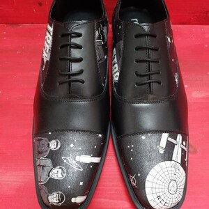 May include: A pair of black leather shoes with a white design featuring characters from Star Trek. The shoes have a lace-up closure and a rounded toe.