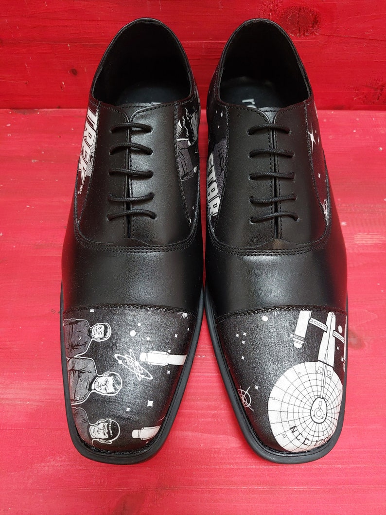 May include: A pair of black leather shoes with a white design featuring characters from Star Trek. The shoes have a lace-up closure and a rounded toe.