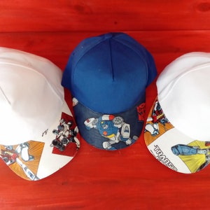 May include: Three baseball caps with a Transformers theme. The caps are white, blue, and white with a colorful Transformers graphic on the front.