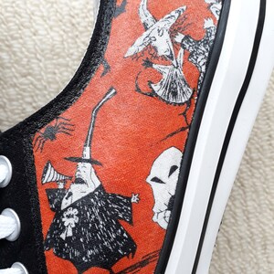 Puede incluir: Un par de zapatos de lona negros con suela blanca y tela naranja con ilustraciones en blanco y negro de personajes de Pesadilla Antes de Navidad. Los personajes incluyen a Jack Skellington, Sally y Oogie Boogie.