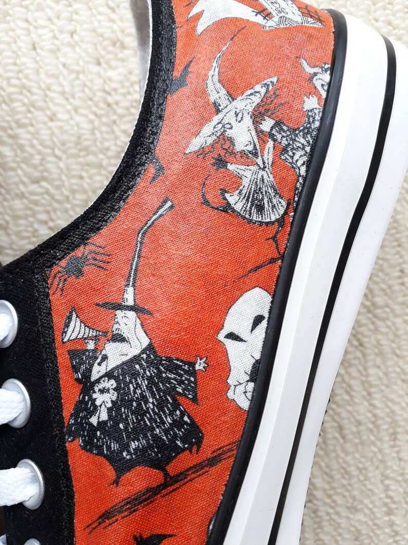 Puede incluir: Un par de zapatos de lona negros con suela blanca y tela naranja con ilustraciones en blanco y negro de personajes de Pesadilla Antes de Navidad. Los personajes incluyen a Jack Skellington, Sally y Oogie Boogie.