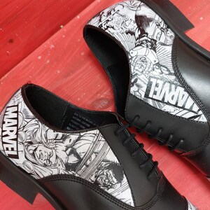 May include: A pair of black leather lace-up shoes with a white and black comic book print featuring Marvel characters. The shoes have a classic oxford style and are decorated with the word "MARVEL" on the side.