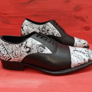May include: A pair of black leather oxford shoes with a white and black comic book print featuring Marvel characters. The shoes have a lace-up closure and a cap toe.