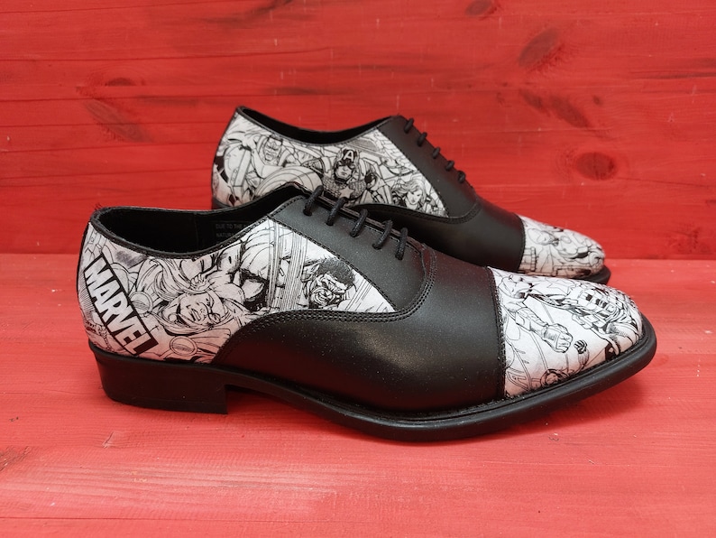 May include: A pair of black leather oxford shoes with a white and black comic book print featuring Marvel characters. The shoes have a lace-up closure and a cap toe.