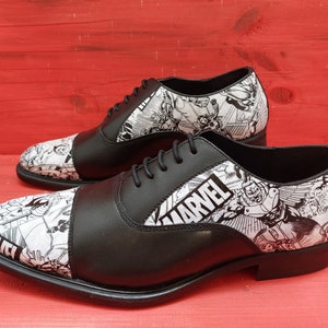 May include: Black leather oxford shoes with a white and black comic book print featuring Marvel characters. The shoes have a cap toe and lace-up closure.