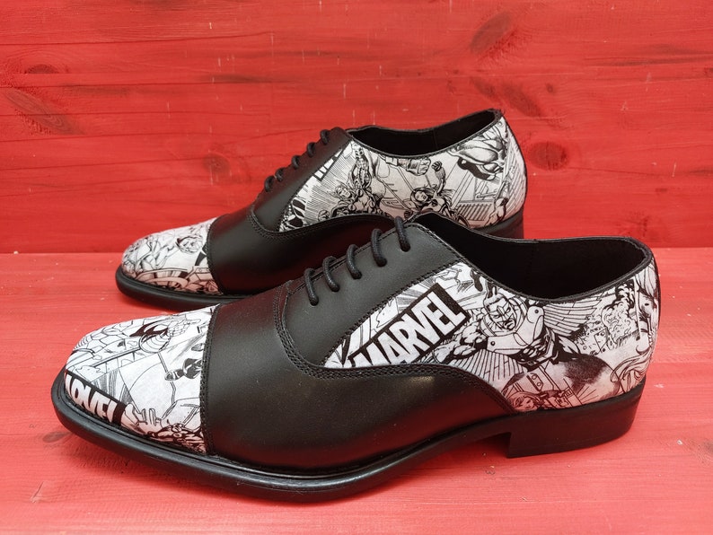 May include: Black leather oxford shoes with a white and black comic book print featuring Marvel characters. The shoes have a cap toe and lace-up closure.
