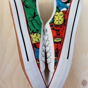 May include: White canvas sneakers with a comic book design featuring Iron Man, Hulk, and Captain America. The sneakers have black laces and a white rubber sole.