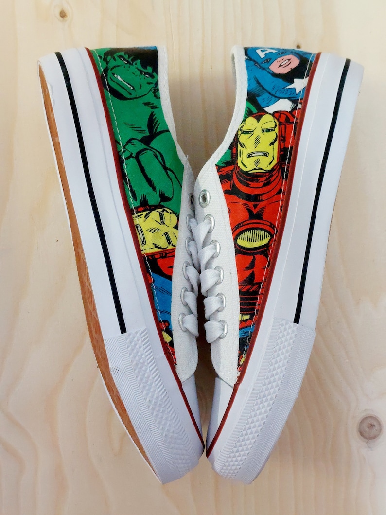 May include: White canvas sneakers with a comic book design featuring Iron Man, Hulk, and Captain America. The sneakers have black laces and a white rubber sole.