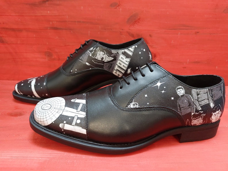 May include: A pair of black leather Oxford shoes with a white and black Star Trek themed design. The shoes feature images of the Star Trek crew and the Enterprise spaceship.