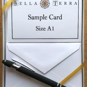 May include: A white card with a black border and the text "Bella Terra" with a compass logo in the center. The text "Sample Card" and "Size A1" are below the logo. The card is in a white box with a gold ribbon and a black pen.
