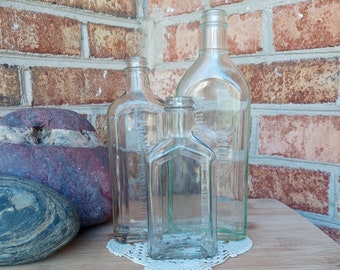 May include: Three clear glass bottles of various shapes and sizes on a wooden surface with a white doily. The bottles are empty and have a vintage look.
