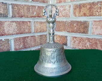 May include: A silver bell with a decorative handle featuring a stylized figure. The bell has intricate carvings and a rounded shape.