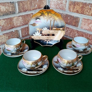 May include: A set of five Japanese teacups and saucers with a black and gold floral design. The cups and saucers feature a mountain, bamboo, and lotus flower design. The set also includes a decorative plate with a similar design.