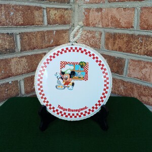 May include: A white ceramic trivet with a red and white checkered border. Mickey Mouse is dressed as a chef and is cooking eggs in a pan. The trivet says "Tokyo Disneyland."