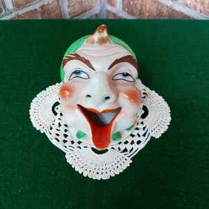May include: A white ceramic figurine of a clown with a green hat, red lips, and a wide smile. The figurine is sitting on a white crocheted doily.