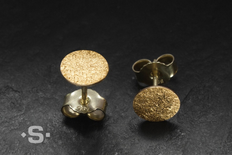 May include: A pair of gold-toned stud earrings with a textured circular design. The earrings are made of metal and have a post-back closure.