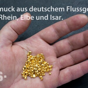 May include: A hand holding a pile of small, shiny gold nuggets. The text "Schmuck aus deutschem Flussgold aus Rhein, Elbe und Isar." is visible above the hand.